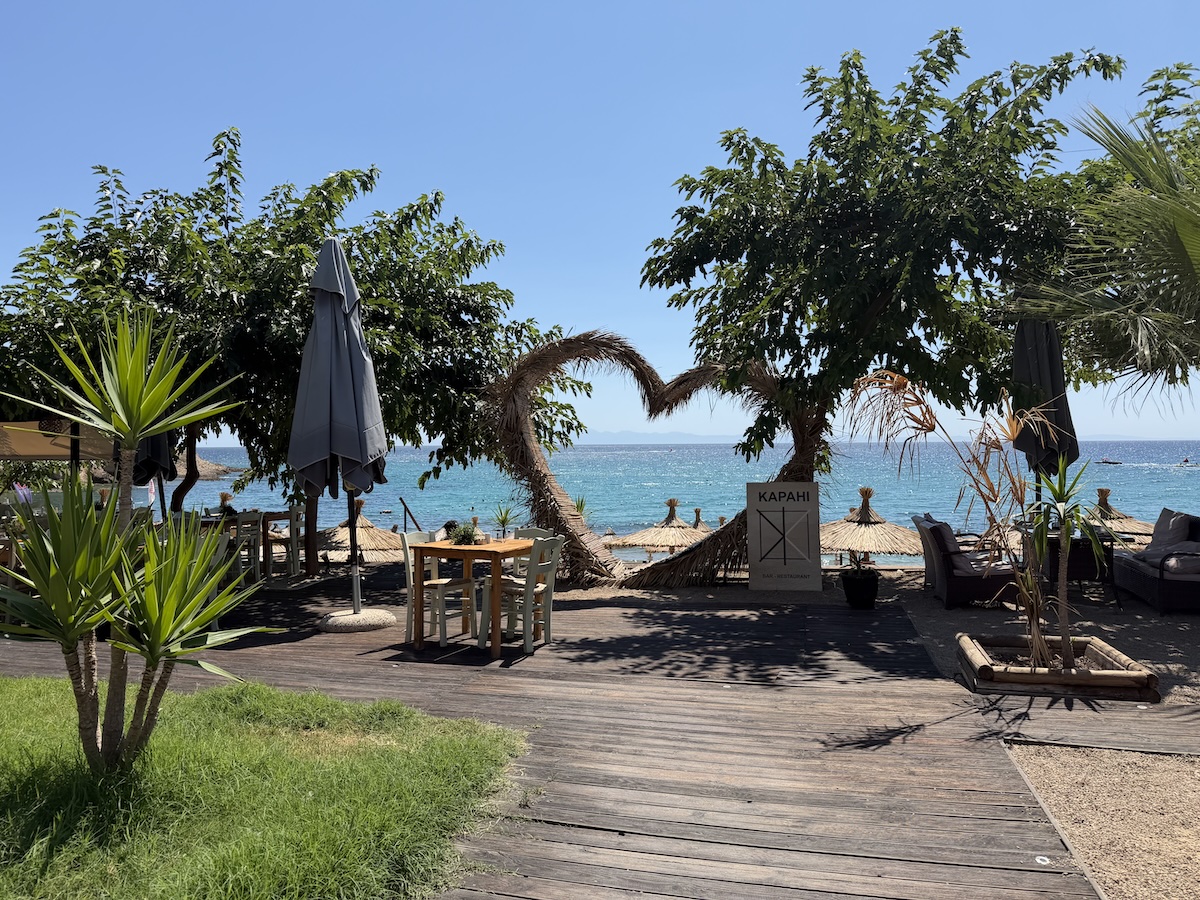 Ruhiger Strandbereich bei Potos auf Thassos mit Holzsteg, Palmen, Liegen und türkisfarbenem Meer unter blauem Himmel.