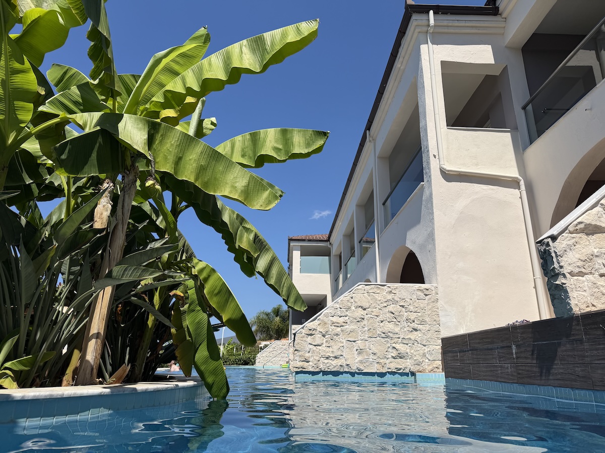 Blaues Poolwasser vor einer Unterkunft bei Potos auf Thassos, umgeben von grünen Bananenblättern und klarem Himmel.