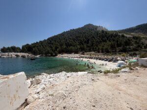 Türkisblaues Wasser, heller Kiesstrand und bewaldete Hügel an der Küste von Thassos bei einem beliebten Badeort