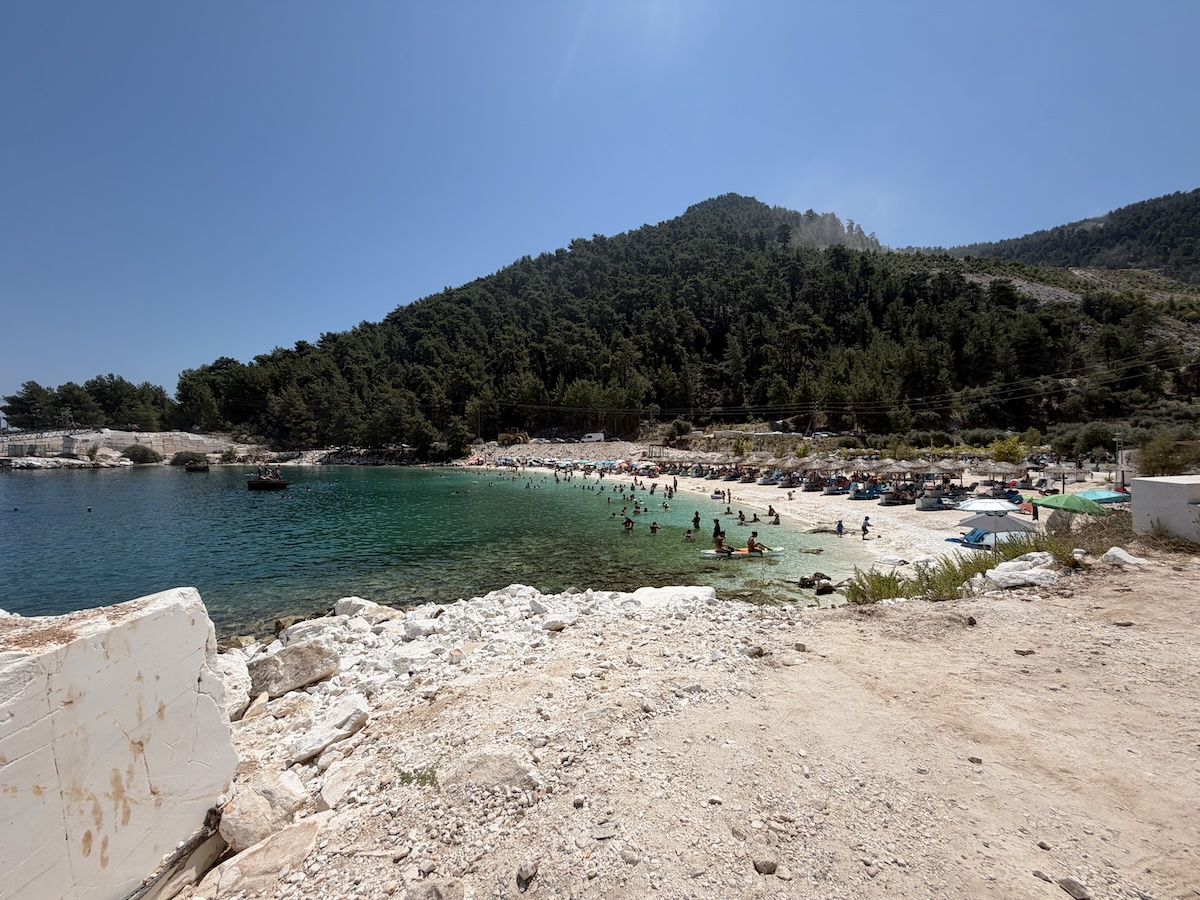 Türkisblaues Wasser, heller Kiesstrand und bewaldete Hügel an der Küste von Thassos bei einem beliebten Badeort