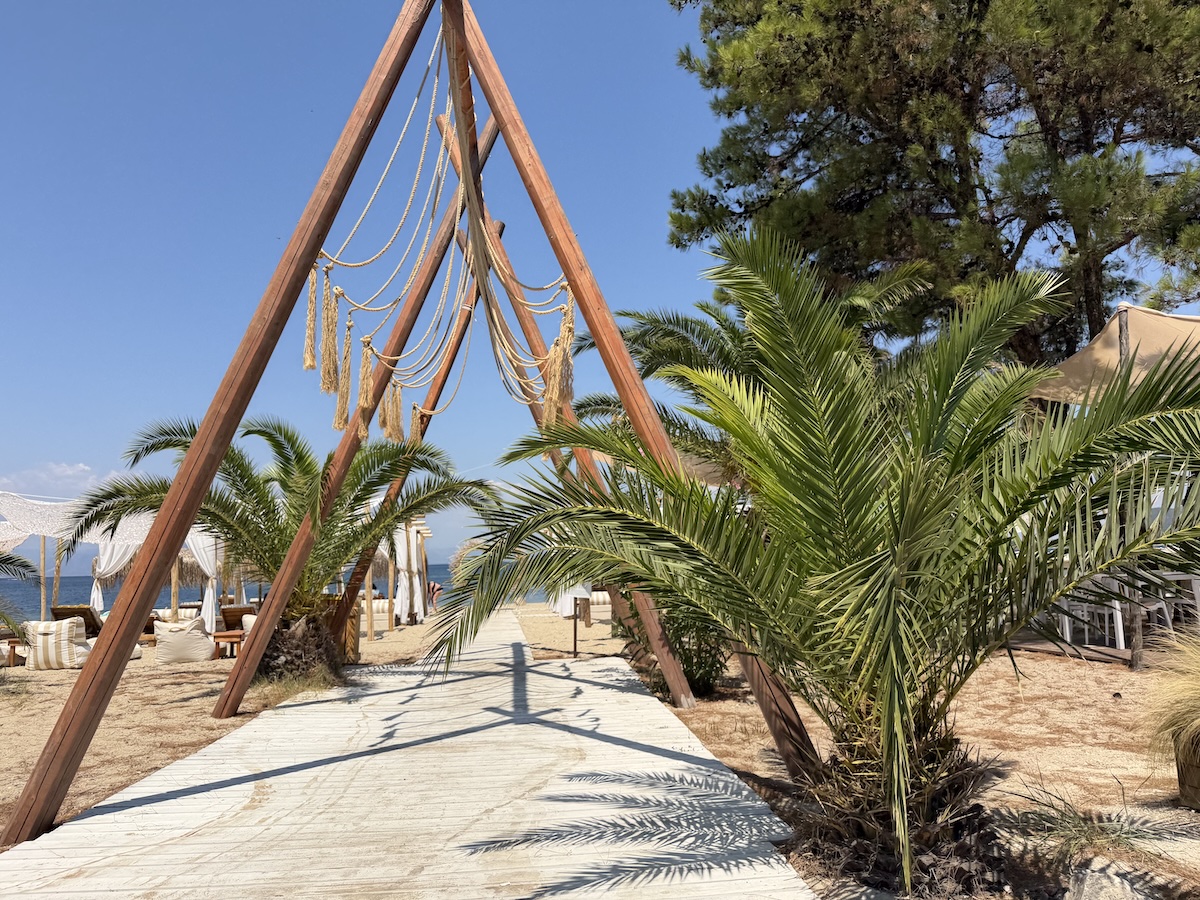 Ein heller Strandweg in Skala Prinos auf Thassos führt zwischen Palmen und einer großen Holzschaukel zum Meer.