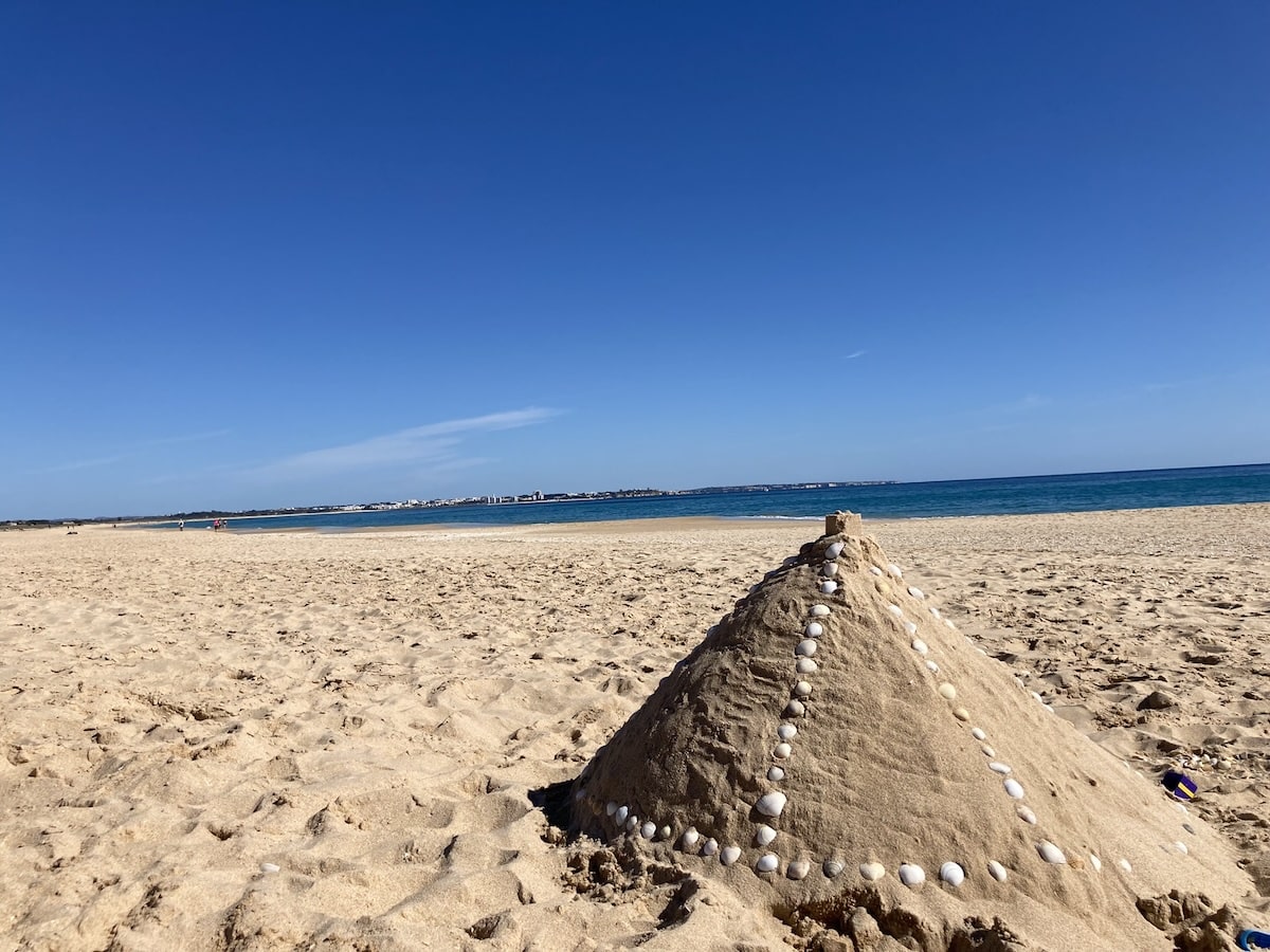 Am breiten Sandstrand von Lagos steht eine mit Muscheln verzierte Sandpyramide, dahinter liegen Meer und ein klarer Himmel.