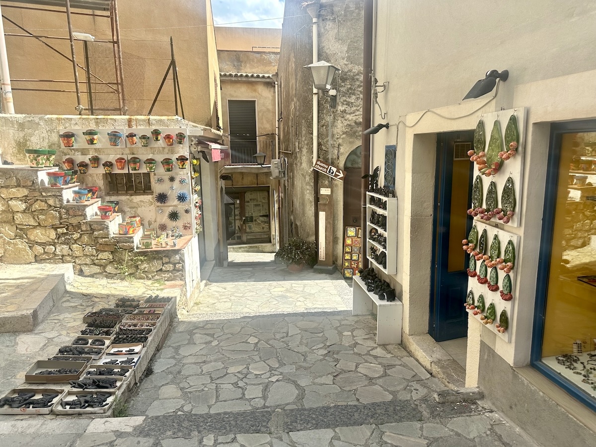 In einer schmalen Altstadtgasse in Castelmola sind bunte Keramikschalen, kleine Figuren und weitere Waren vor den Häusern ausgestellt.