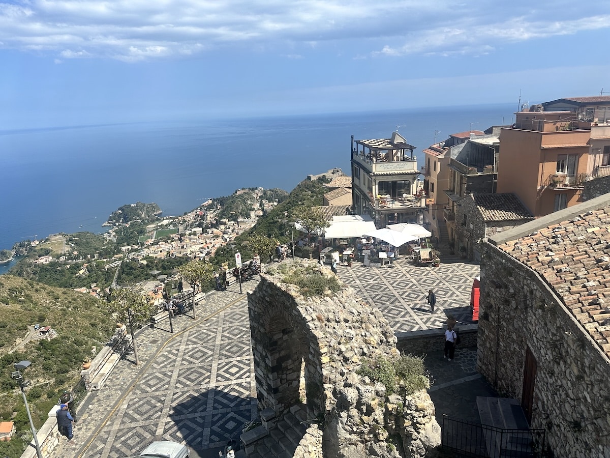 Ein gepflasterter Platz über einer Küstenstadt blickt auf das offene Meer, umgeben von Häusern und Felsen.