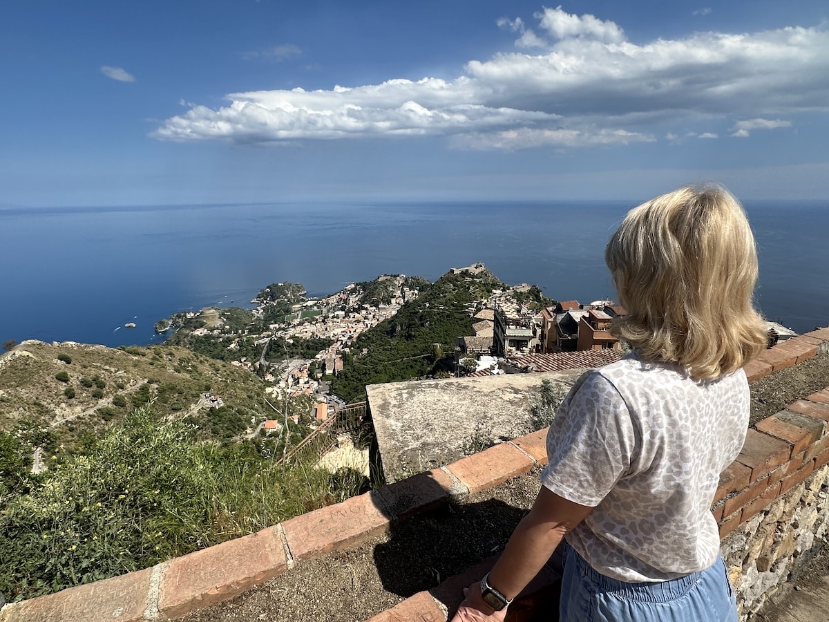 Eine blonde Frau blickt von einer Aussichtsterrasse von Castelmola über Taormina auf das tiefblaue Meer und die Küste.