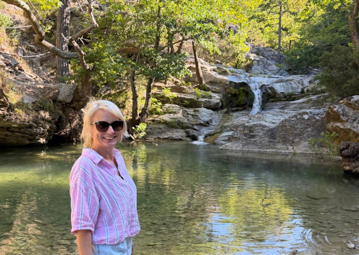 Mandy steht mit rosa Hemd an einem natürlichen Badebecken unter einem Wasserfall auf Thassos in Griechenland.