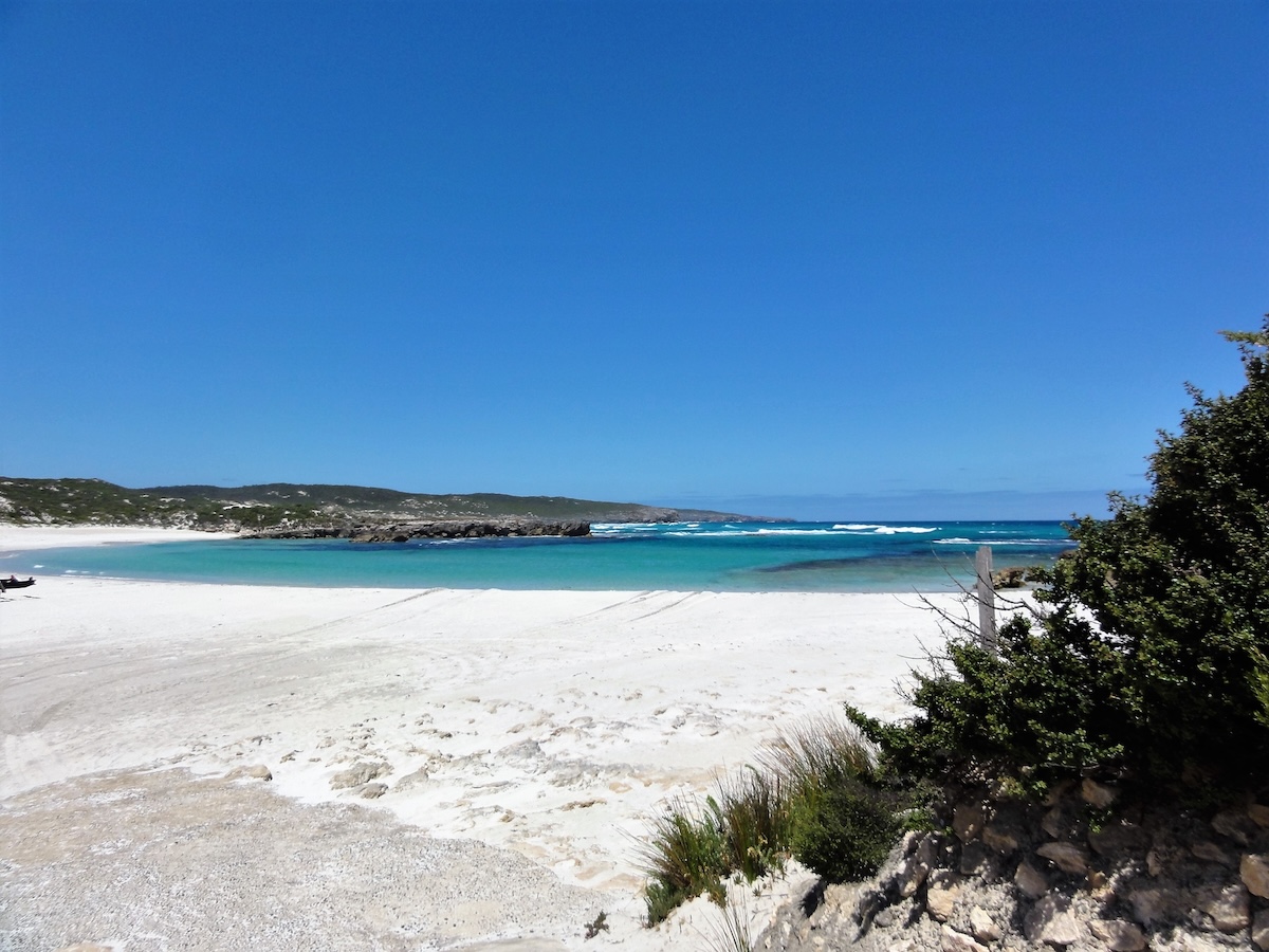 Weißer Sandstrand an einer türkisfarbenen Lagune mit felsiger Küste, flachen Wellen und dichtem Küstenbewuchs