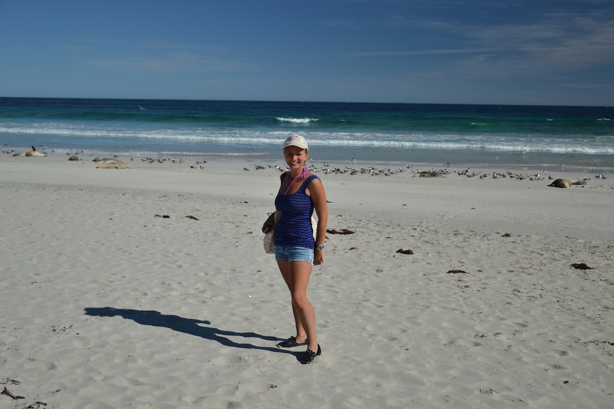 Eine lächelnde Reisende im blauen Oberteil steht am weißen Strand vor Wellen, Robben und zahlreichen Möwen.