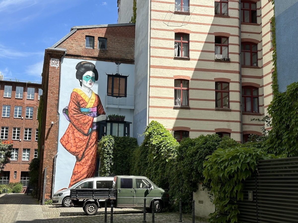 Ein Berliner Streetart-Wandgemälde zeigt eine bunt gekleidete Frau zwischen Backsteinhäusern und überwucherten Mauern.