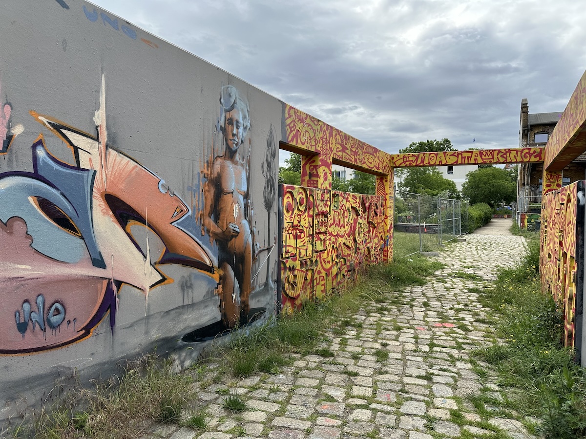 Bunte Berliner Streetart mit großflächigen Graffiti, einer Figur und Murals entlang eines überwachsenen Kopfsteinpflasterwegs