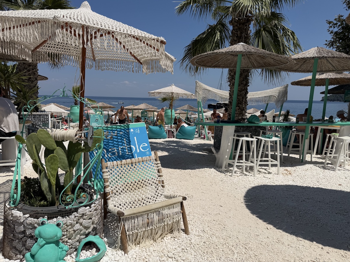Weiße Kiesel, geflochtene Sonnenschirme und Palmen prägen die Strandbar am Marble Beach auf Thassos, direkt am blauen Meer.