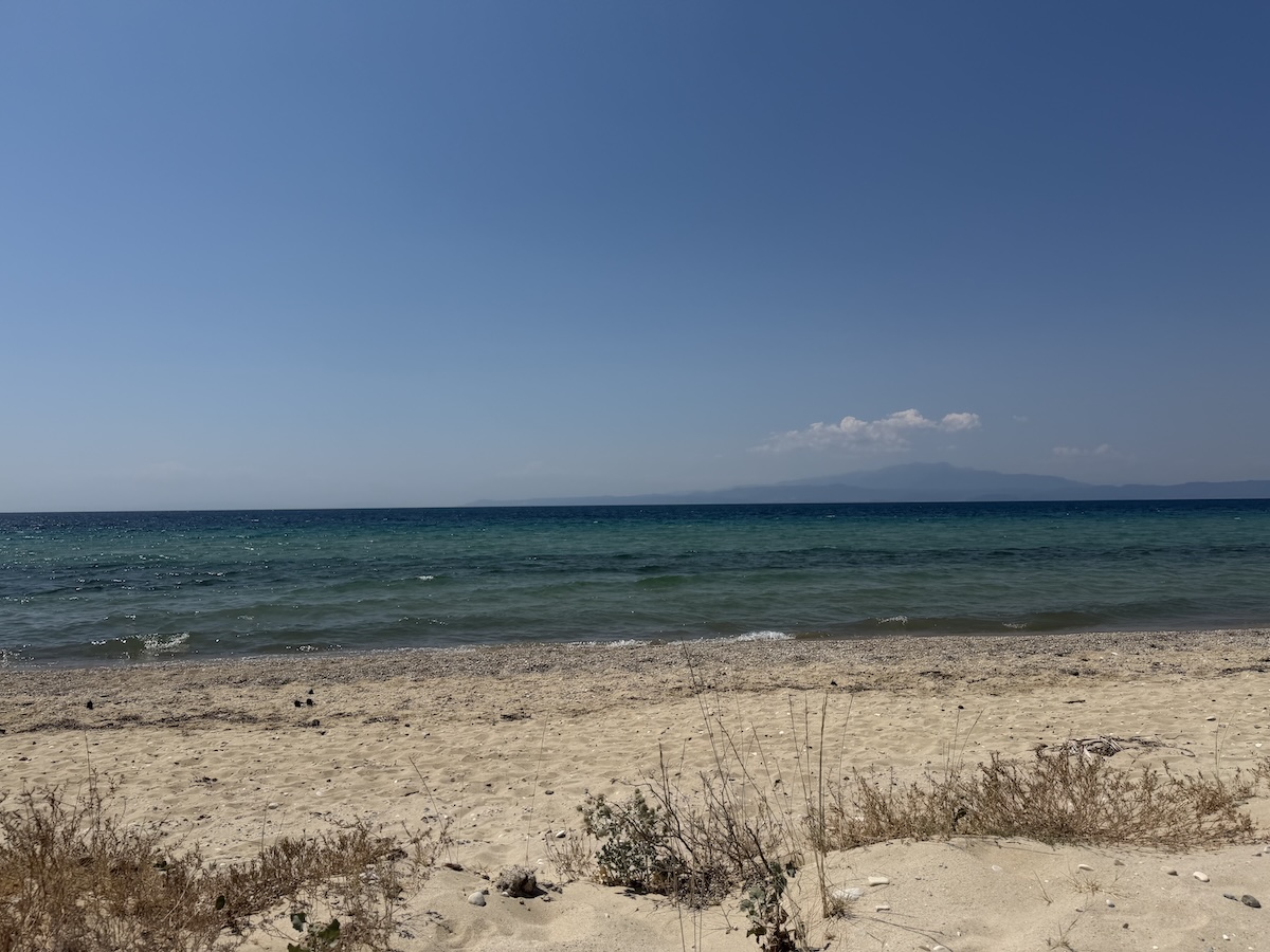 Am sandigen Strand von Thassos schimmert türkisgrünes Meer; am Horizont zeichnet sich eine Küste unter klarem Himmel ab.