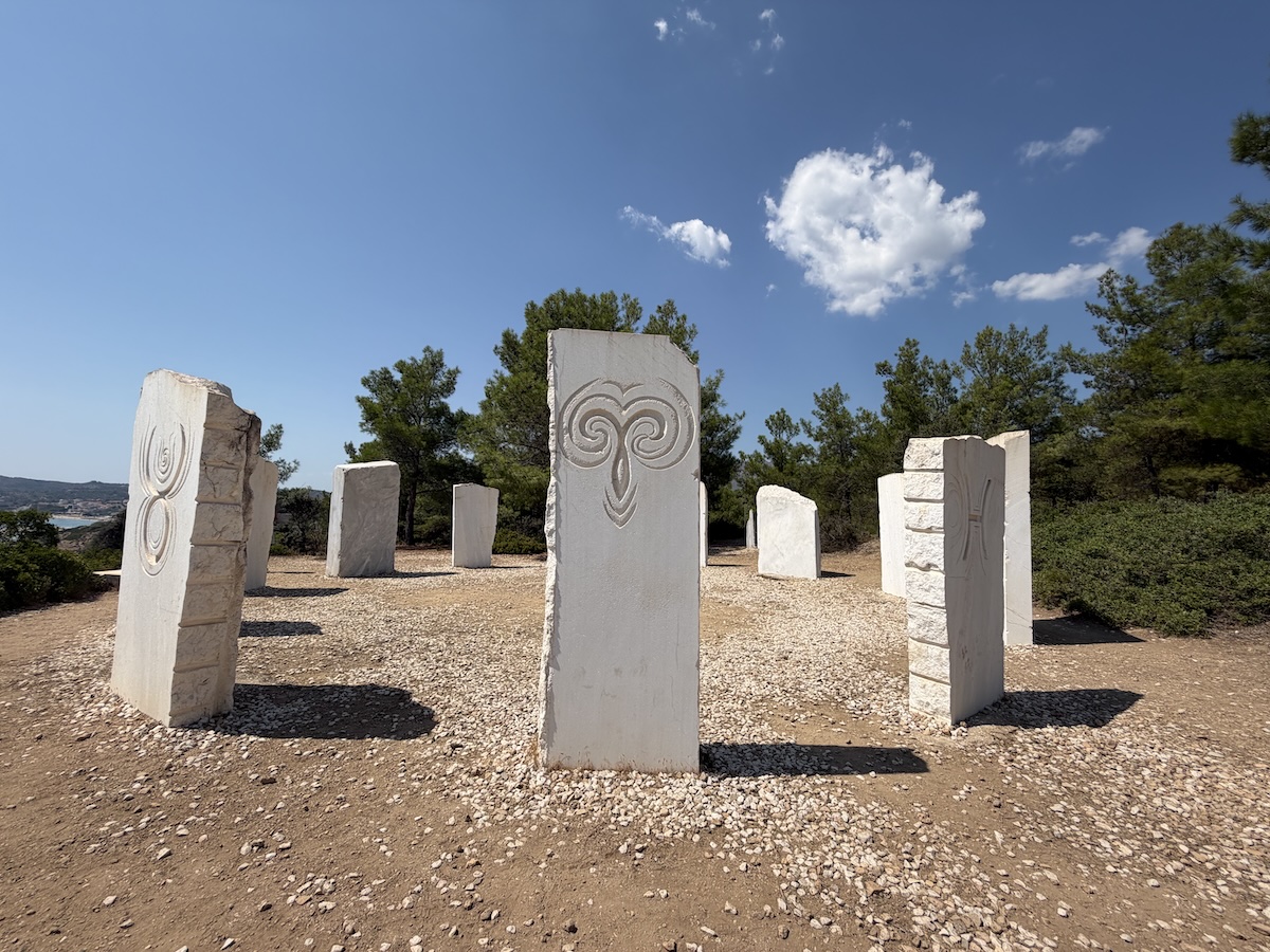 Auf Thassos stehen kunstvoll reliefierte weiße Marmorstelen auf einem steinigen Platz, umgeben von Pinien und blauem Himmel.