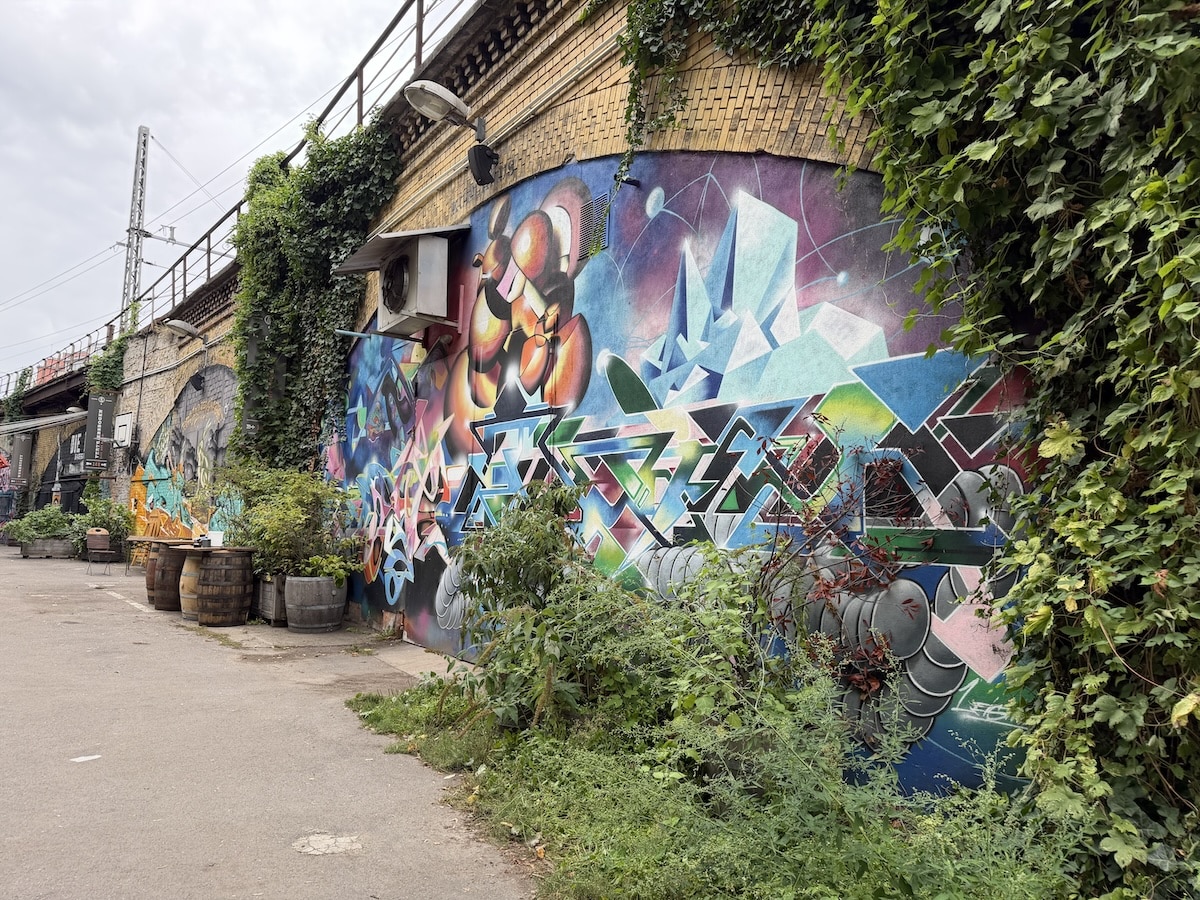 Buntes Streetart-Mural mit geometrischen Formen schmückt eine Backsteinwand in Berlin zwischen Pflanzen und alten Fässern.