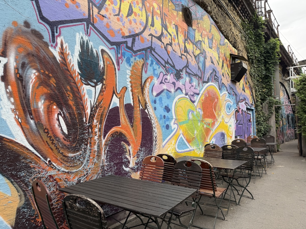 Bunte Streetart mit abstrakten Formen und Figuren schmückt eine Berliner Hauswand neben einem Außenbereich mit Tischen.