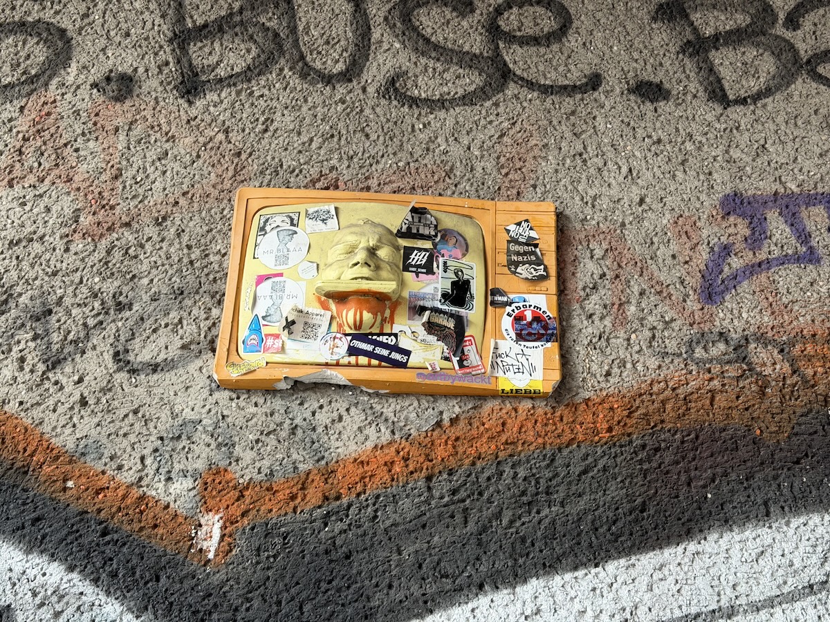 Auf einer rauen Betonfläche liegen bunte Sticker und Paste-ups neben orangefarbenen und schwarzen Graffiti in Berlin.