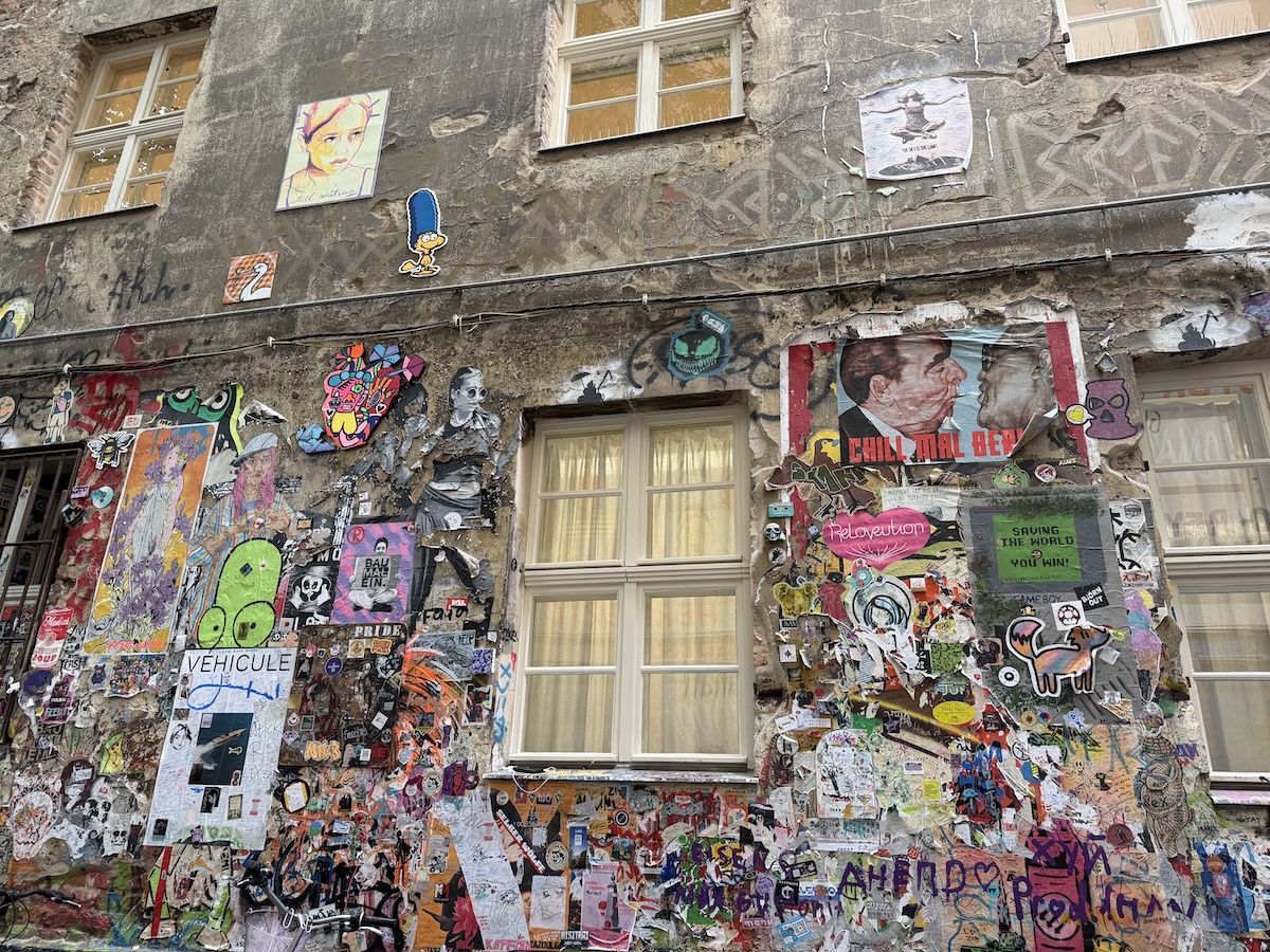 Bunte Streetart mit Murals, Paste-ups und Stickern bedeckt eine verwitterte Berliner Hausfassade nahe verschiedener Kieze.