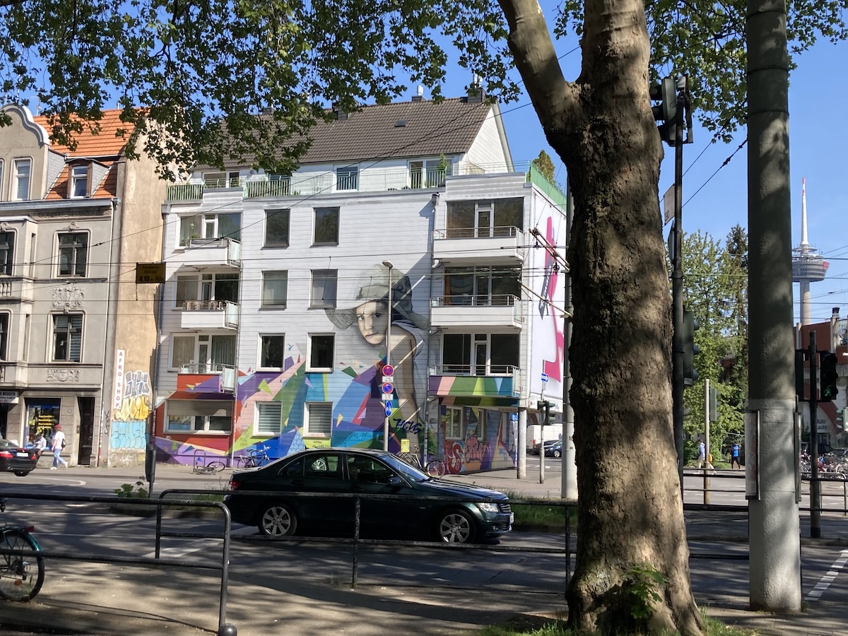 Farbenfrohe Streetart-Murals schmücken ein Wohnhaus an einer Straße in Köln-Ehrenfeld neben Bäumen und Straßenbahnmasten.