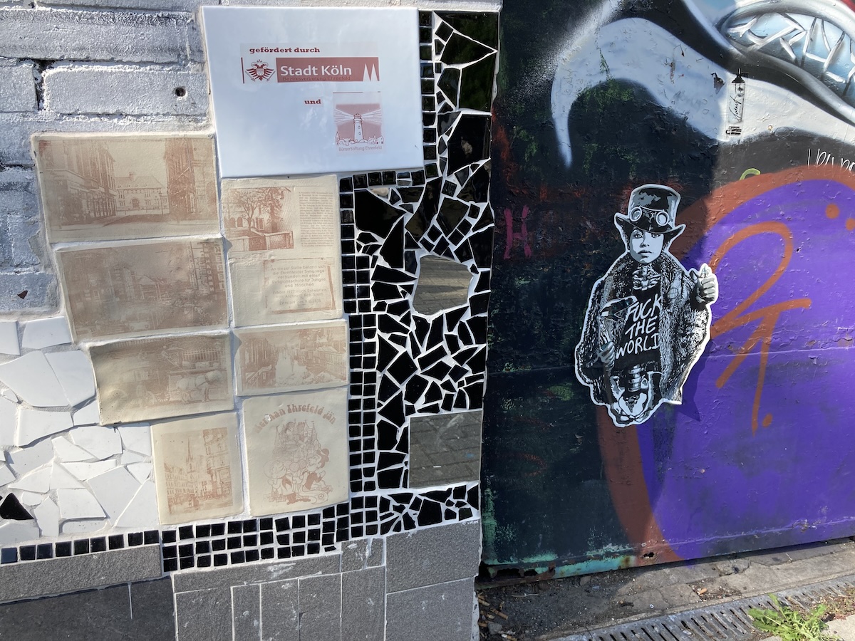 Mosaik und Streetart mit Porträt eines Kindes an einer Hauswand ergänzt durch ein Schild der Stadt Köln.