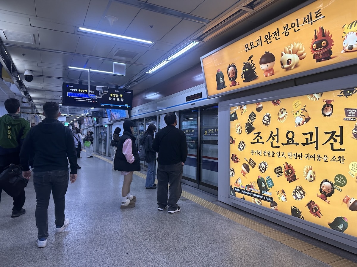Fahrgäste warten an einem Bahnsteig der Seoul Metro neben gelben koreanischen Werbetafeln und einem Zugzugang