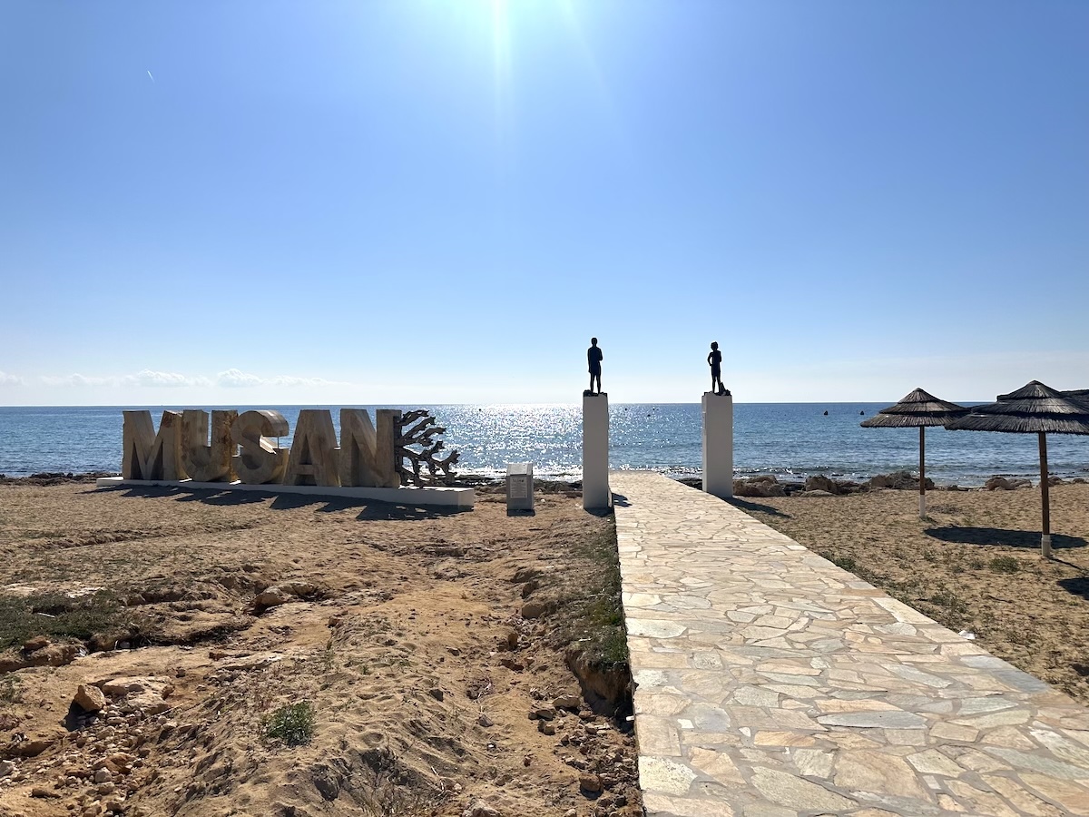 Sandige Küste bei Ayia Napa mit steinernem Weg, Sonnenschirmen und der MUSEAN-Skulptur am glitzernden Meer