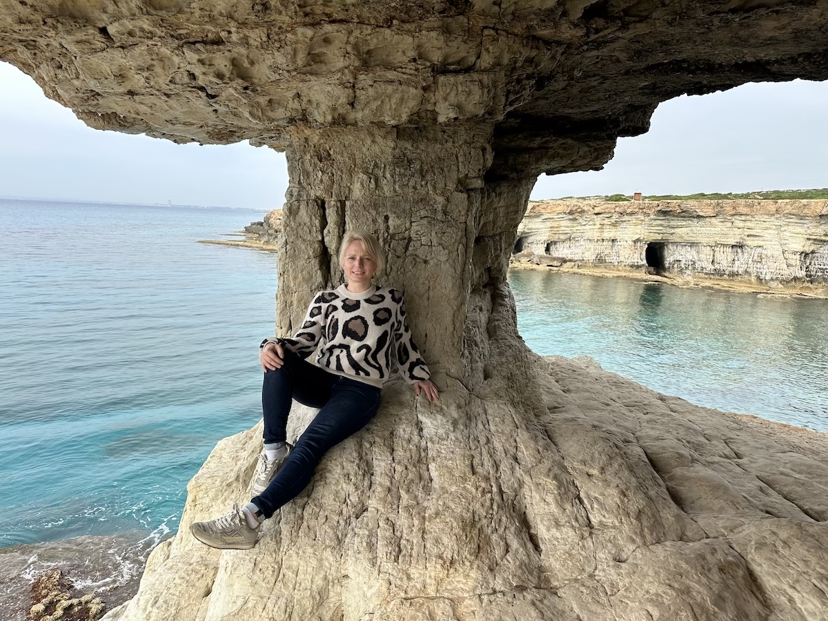 Eine Reisende sitzt unter einem Felsbogen an den Sea Caves bei Cape Greco nahe Ayia Napa am türkisfarbenen Meer.
