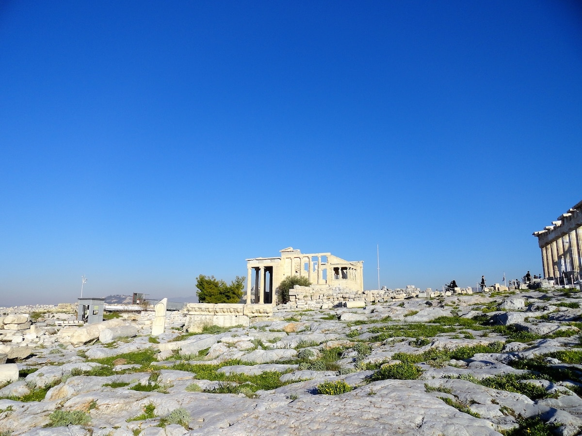 Antike Ruinen auf dem felsigen Gelände der Akropolis in Athen, unter strahlend blauem Himmel und mit Blick über die Stadt.
