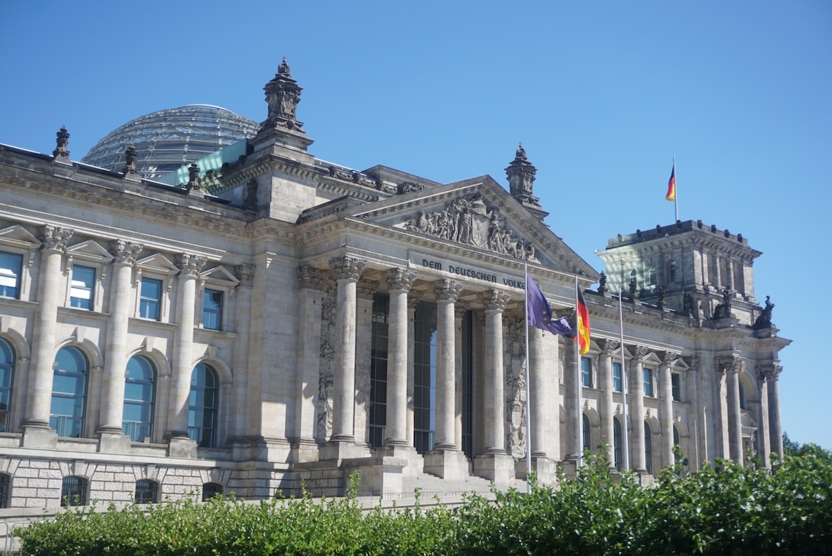 Vor dem blauen Himmel zeigt sich das Berliner Reichstagsgebäude mit seiner Glaskuppel, dem Säulenportal und wehenden Flaggen.